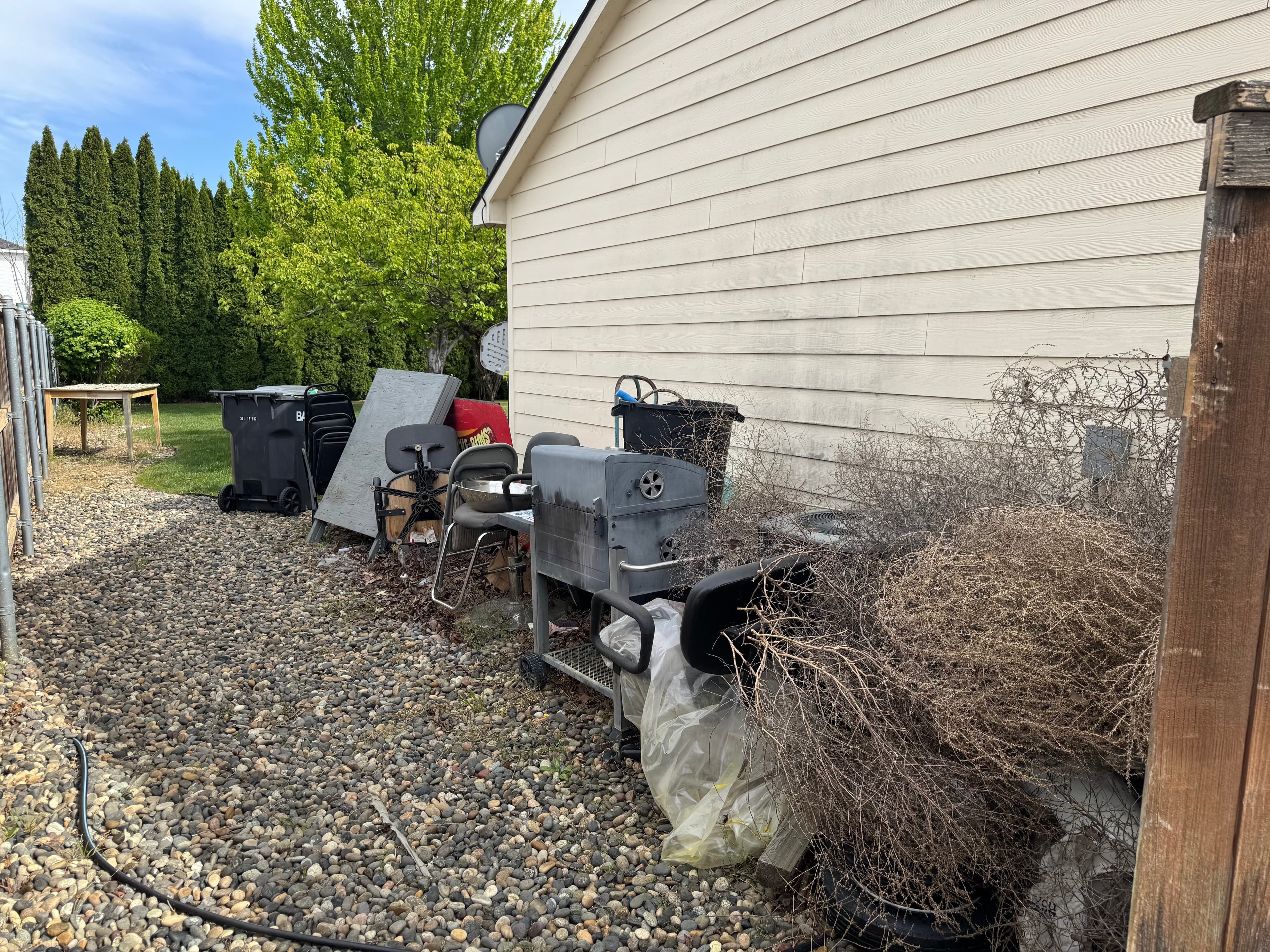 Cluttered side yard before junk removal with outdoor furniture, pool toys, bins, panels, chairs, and debris